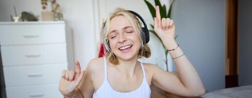 portret van gelukkig en zorgeloos blond vrouw, luistert naar muziek- in draadloze koptelefoon, dansen en het zingen langs favoriete liedje, uitgeven tijd alleen Bij huis foto