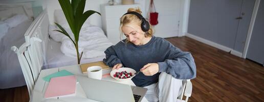 portret van glimlachen jong vrouw, aan het kijken TV tonen in koptelefoon, aan het eten ontbijt en op zoek Bij laptop scherm foto