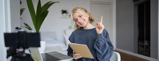 portret van jong lachend, charismatisch vrouw, verhogen een vinger, onderwijs online, lezing van notitieboekje, op zoek Bij laptop, opname Cursus, inhoud maker werken Aan nieuw project van huis foto