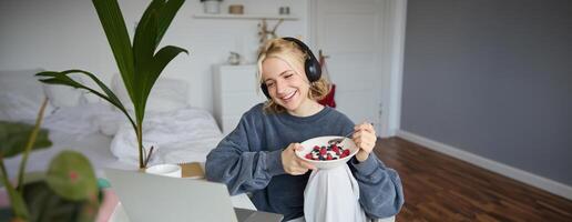 portret van glimlachen mooi vrouw, zittend in kamer met ontbijt, aan het eten en aan het kijken TV tonen Aan laptop, lachend en op zoek Bij scherm foto