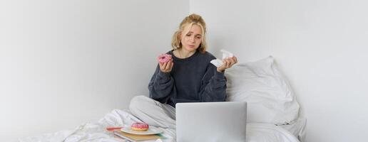 portret van verdrietig, huilen jong vrouw, blijven Bij huis, zittend in bed met donut en comfort voedsel, op zoek Bij iets verontrustend Aan laptop scherm foto