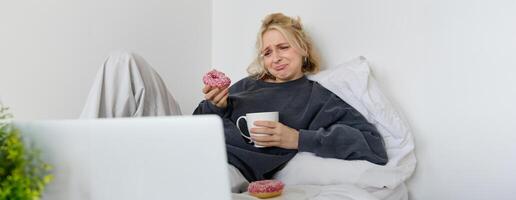 portret van verdrietig, huilen jong vrouw, blijven Bij huis, zittend in bed met donut en comfort voedsel, op zoek Bij iets verontrustend Aan laptop scherm foto