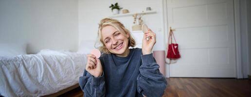 portret van jong vrouw, inhoud Schepper, tonen schoonheid bedenken producten Bij camera, zittend Aan verdieping in slaapkamer en glimlachen foto