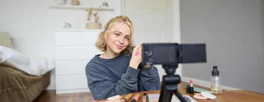 beeld van jong elegant vrouw, blogger opname een schoonheid levensstijl van haar plukken het beste lippenstift, tonen lip balsem stalen Aan haar huid, zittend in voorkant van digitaal camera in leeg kamer foto