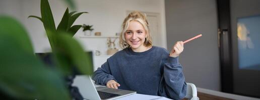 portret van jong charismatisch vrouw, lachend, Holding pen, op zoek Bij laptop, chatten, aan het doen online Cursus, beantwoorden een vraag, aan het studeren met tutor foto