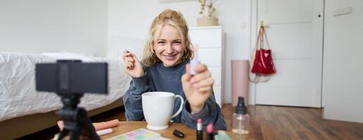 levensstijl, schoonheid blogger, vrouw opname van haar zetten Aan verzinnen, pratend naar camera, maken online zelfstudie, tonen haar lip glans of lippenstift naar volgers, zittend Aan verdieping met kop van thee foto