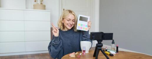 portret van mooi levensstijl blogger, meisje records een Aan haar camera voor sociaal media, shows palet van oogschaduw, doet een bedenken zelfstudie voor haar volgers, zit in haar kamer foto