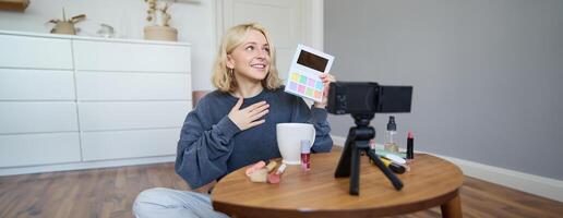 portret van mooi levensstijl blogger, meisje records een Aan haar camera voor sociaal media, shows palet van oogschaduw, doet een bedenken zelfstudie voor haar volgers, zit in haar kamer foto