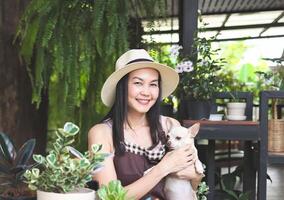 Aziatisch vrouw tuinman vervelend hoed en schort zittend Bij tafel met fabriek potten en tuinieren hulpmiddelen, Holding bruin chihuahua hond, glimlachen en op zoek Bij camera. foto