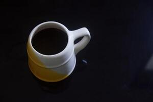 zwart koffie in een wit en geel kop Aan een zwart keramisch tafel foto