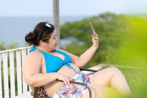 Aziatisch plus zo vrouw vervelend bikini gebruik makend van mobiel telefoon naar selfie haarzelf terwijl vakantie Bij zee in zomer. concept van positief lichaam figuur. foto
