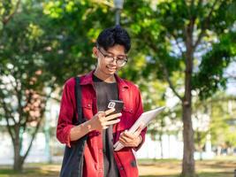 jong Aziatisch college leerling gebruik makend van smartphone met gelukkig uitdrukking. een mannetje glimlachen terwijl Holding zijn telefoon en boeken Bij de openbaar park. kopiëren ruimte foto