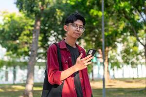 jong Aziatisch college leerling gebruik makend van smartphone met gelukkig uitdrukking. een mannetje glimlachen terwijl Holding zijn telefoon en boeken Bij de openbaar park. kopiëren ruimte foto