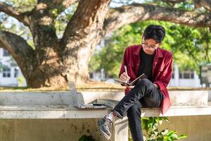 Aziatisch college leerling aan het studeren Aan laptop Bij campus buitenshuis park. Mens schrijven Aan een Notitie boek en werken Aan laptop. leerzaam concept foto