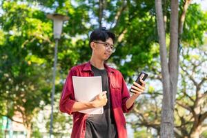 jong Aziatisch college leerling gebruik makend van smartphone met gelukkig uitdrukking. een mannetje glimlachen terwijl Holding zijn telefoon en boeken Bij de openbaar park. kopiëren ruimte foto