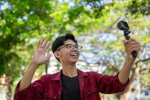 Aziatisch Mens opname en pratend naar camera. jong Mens vloggen en nemen selfie met actie camera Bij openbaar park. foto