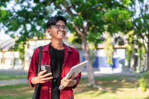 jong Aziatisch college leerling gebruik makend van smartphone met gelukkig uitdrukking. een mannetje glimlachen terwijl Holding zijn telefoon en boeken Bij de openbaar park. kopiëren ruimte foto