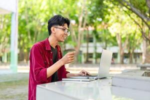 jong Aziatisch Mens glimlachen terwijl gebruik makend van laptop voor werken en conferentie vergadering Bij de ochtend. Aziatisch Mens hebben een telefoontje Aan laptop. concept van afgelegen en freelance foto