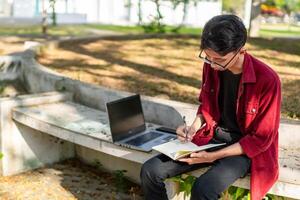 Aziatisch college leerling aan het studeren Aan laptop Bij campus buitenshuis park. Mens schrijven Aan een Notitie boek en werken Aan laptop. leerzaam concept foto