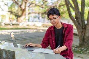 Aziatisch college leerling aan het studeren Aan laptop Bij campus buitenshuis park. Mens schrijven Aan een Notitie boek en werken Aan laptop. leerzaam concept foto