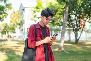 jong Aziatisch college leerling gebruik makend van smartphone met gelukkig uitdrukking. een mannetje glimlachen terwijl Holding zijn telefoon en boeken Bij de openbaar park. kopiëren ruimte foto