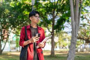 jong Aziatisch college leerling gebruik makend van smartphone met gelukkig uitdrukking. een mannetje glimlachen terwijl Holding zijn telefoon en boeken Bij de openbaar park. kopiëren ruimte foto