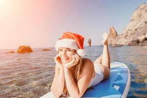 vrouw zee sup. dichtbij omhoog portret van gelukkig jong Kaukasisch vrouw met lang haar- in de kerstman hoed op zoek Bij camera en lachend. schattig vrouw portret in een wit bikini poseren Aan sup bord in de zee foto