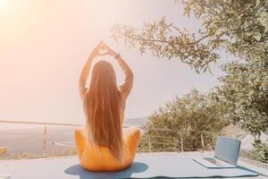 geschiktheid vrouw zee. gelukkig midden- oud vrouw in oranje sportkleding opdrachten ochtend- buitenshuis Aan yoga mat met laptop in park over- oceaan strand. vrouw geschiktheid pilates yoga routine. gezond levensstijl. foto