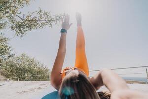 geschiktheid vrouw zee. gelukkig midden- oud vrouw in oranje sportkleding opdrachten ochtend- buitenshuis Aan yoga mat met laptop in park over- oceaan strand. vrouw geschiktheid pilates yoga routine. gezond levensstijl. foto