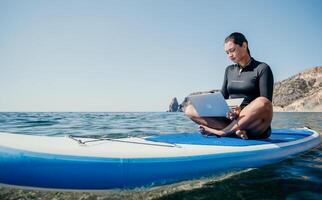 vrouw zee laptop. digitaal nomade, freelancer met laptop werken Aan sup bord Bij kalmte zee strand. gelukkig glimlachen meisje verlicht spanning van werk. freelance, digitaal nomade, reizen en vakantie concept foto