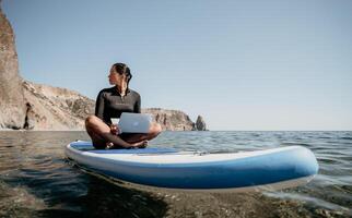 vrouw zee laptop. digitaal nomade, freelancer met laptop werken Aan sup bord Bij kalmte zee strand. gelukkig glimlachen meisje verlicht spanning van werk. freelance, digitaal nomade, reizen en vakantie concept foto