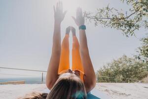 geschiktheid vrouw zee. gelukkig midden- oud vrouw in oranje sportkleding opdrachten ochtend- buitenshuis Aan yoga mat met laptop in park over- oceaan strand. vrouw geschiktheid pilates yoga routine. gezond levensstijl. foto