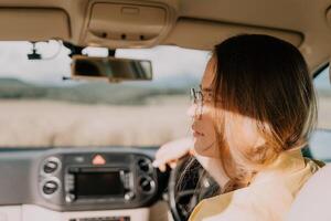 een vrouw in een geel overhemd is het rijden een auto. ze is vervelend bril en heeft haar handen Aan de stuurinrichting wiel foto