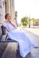 elegant midden- leeftijd vrouw in een wit wijnoogst jurk zittend Aan een bank in de ochtend- stad foto
