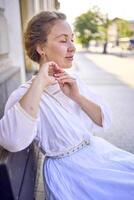 elegant midden- leeftijd vrouw in een wit wijnoogst jurk zittend Aan een bank in de ochtend- stad foto