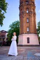 chique jong vrouw in een wit wijnoogst jurk Aan de plein in de buurt de historisch water toren in vinnytsia foto