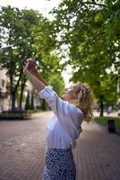 een mooi midden- leeftijd vrouw in jaren 70, 80s stijl kleren strekt zich uit na werk Aan een zon doorweekt steeg foto