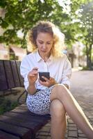 een mooi midden- leeftijd vrouw in jaren 70, 80s stijl kleren is op zoek Bij haar smartphone Aan een bank foto