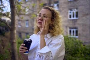 midden- leeftijd vrouw in jaren 70, 80s stijl kleren met een koffie bekladden Aan haar blouse in voorkant van grijs huizen van de jaren 70 foto