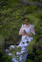 een mooi vrouw in een wit wijnoogst jurk met een trein is bidden terwijl zittend Aan een gedaald boom foto