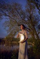 een vrouw in een wit wijnoogst jurk Aan de kust van een meer Bij zonsondergang houdt een maan vormig spiegel foto