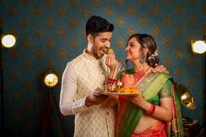 een schattig paar- van man en vrouw in Indisch traditioneel outfits gedurende diwali festival aan het eten laddu terwijl staand in voorkant van de camera foto