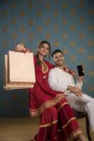 voorraad foto van mooi liefde paar gezeten Aan een stoel geklemd boodschappen doen Tassen en smartphones terwijl gekleed in traditioneel diwali kleding