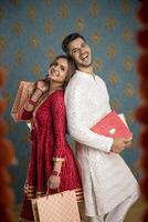afbeelding shows een mooi man en vrouw in traditioneel kleding, grijnzend, geklemd boodschappen doen Tassen en cadeaus voor de diwali viering, en poseren rug aan rug voor de camera foto