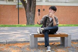 een vrolijk jong Aziatisch Mens in gewoontjes slijtage zit Aan een bank in een park gebruik makend van zijn smartphone. foto