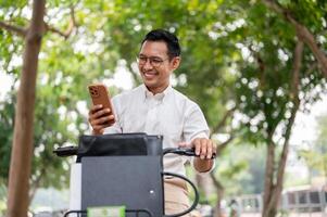 een Vrolijk, zorgeloos Aziatisch zakenman is controle berichten Aan zijn smartphone terwijl hij is Aan een fiets. foto