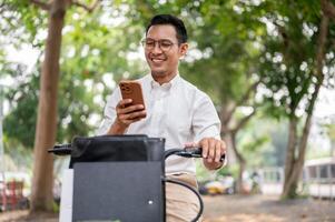 een Vrolijk, zorgeloos Aziatisch zakenman is controle berichten Aan zijn smartphone terwijl hij is Aan een fiets. foto