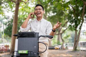 een gelukkig Aziatisch zakenman is pratend Aan de telefoon terwijl hij is Aan een fiets, rijden een fiets naar werk. foto