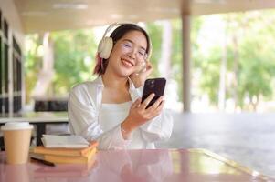 een gelukkig Aziatisch vrouw leerling is genieten van muziek- Aan haar hoofdtelefoons terwijl zittend Bij de cafetaria. foto