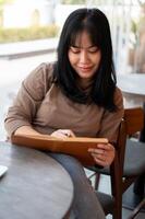 een positief Aziatisch vrouw is houden haar dagboek of lijst haar ideeën in een boek terwijl zittend in een cafe. foto